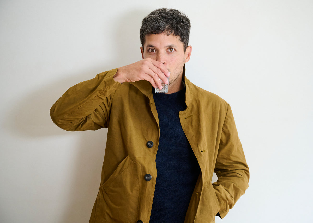A man wearing a tobacco colour dry waxed duster coat, standing against a plain background, drinking from a water glass.