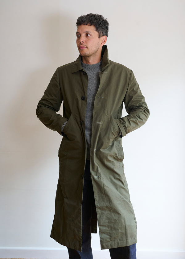 A man wearing a khaki brown dry waxed cotton duster coat with a crew neck sweater underneath. The coat has a full-length design with visible pockets and a buttoned front.