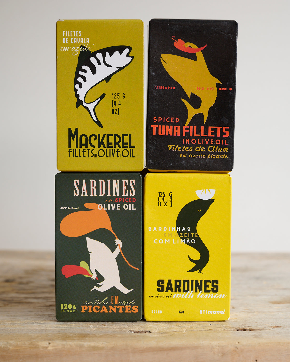 A collection of four tins of canned fish, each with colorful and illustrated labels depicting the type of fish and flavor, arranged on a wooden surface.