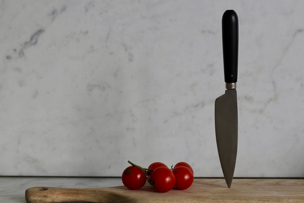 A carbon steel knife with an ebony handle resting on a wooden cutting board, with three small red tomatoes beside it.