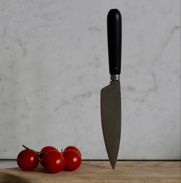 A carbon steel knife with an ebony handle resting on a wooden cutting board, with three small red tomatoes beside it.