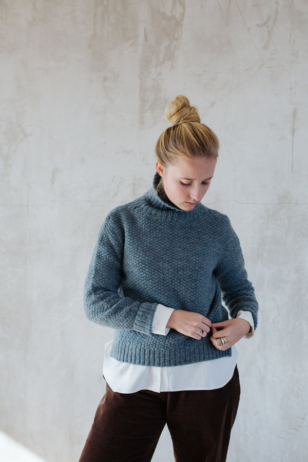 A woman wearing a graphite gray moss stitch lambswool jumper with a funnel neck, slit side detail, and a longer back length.