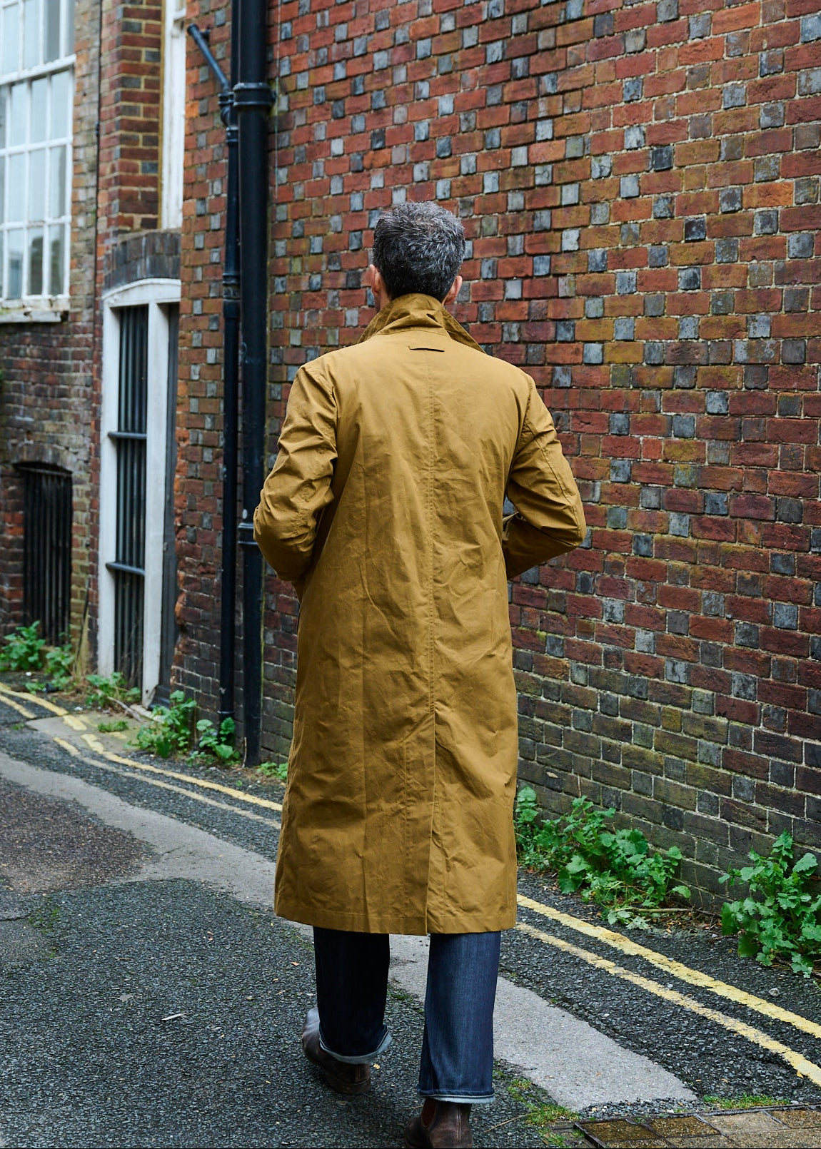 Person wearing a breen colour dry wax coat walking away past a brick wall.