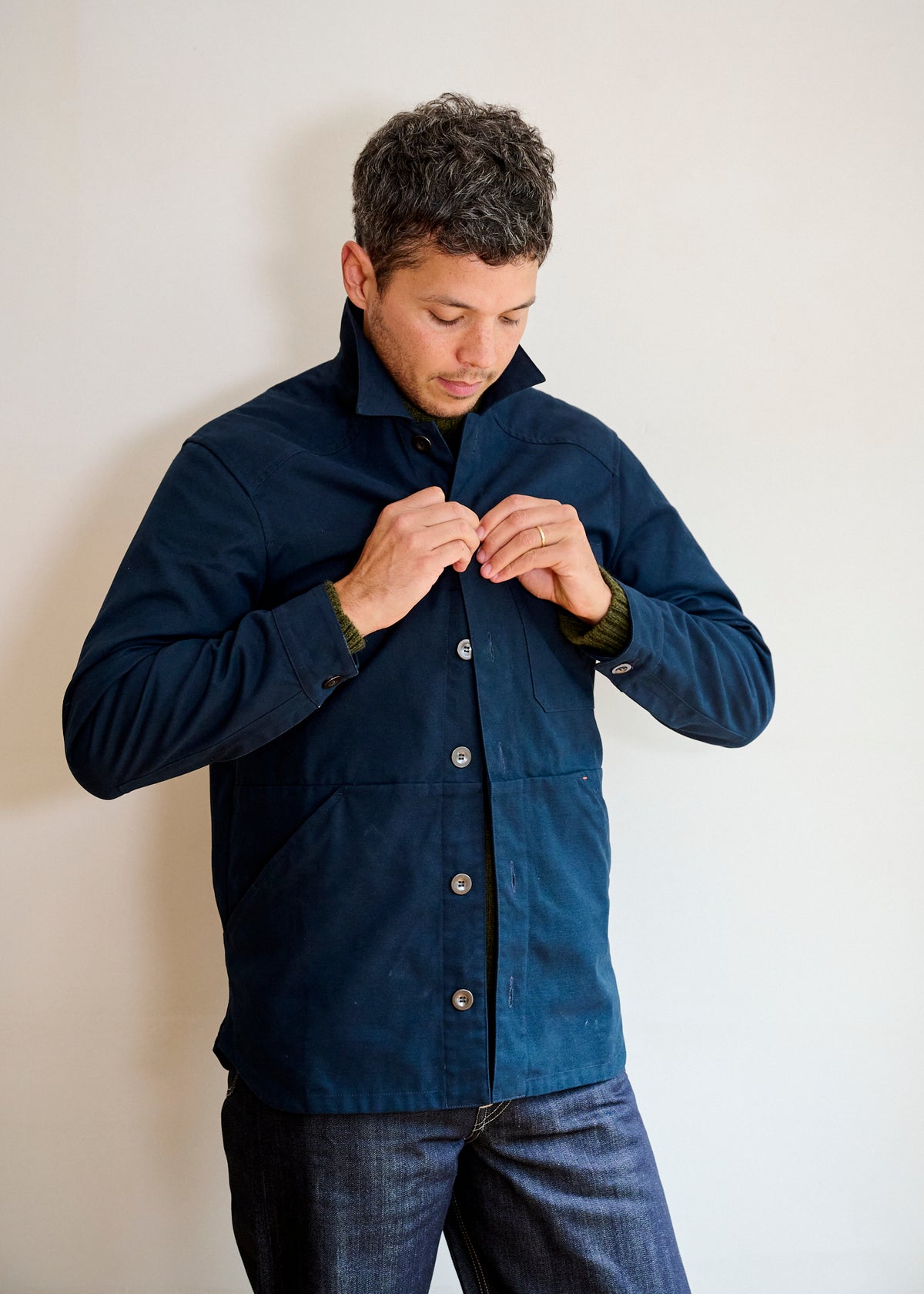 A man wearing a dark navy men's cotton canvas jacket with bronze buttons, paired with jeans, standing against a neutral background.