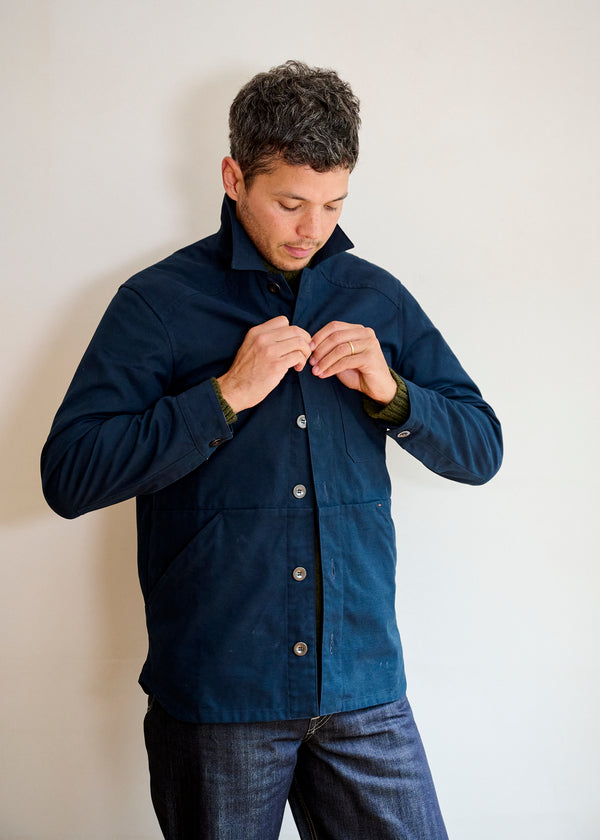 A man wearing a dark navy men's cotton canvas jacket with bronze buttons, paired with jeans, standing against a neutral background.