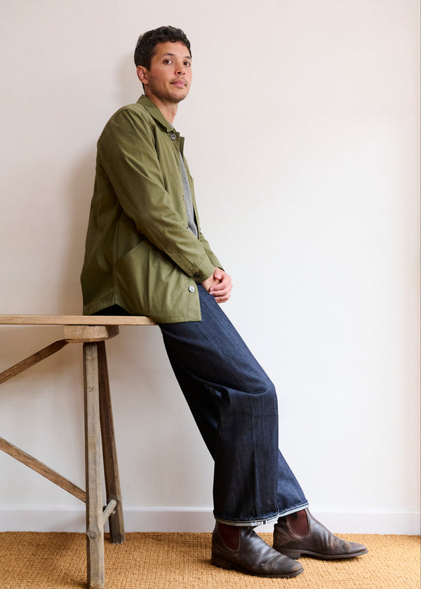 A man sitting on a bench wearing a green men's canvas jacket with bronze buttons, paired with blue jeans and brown shoes.
