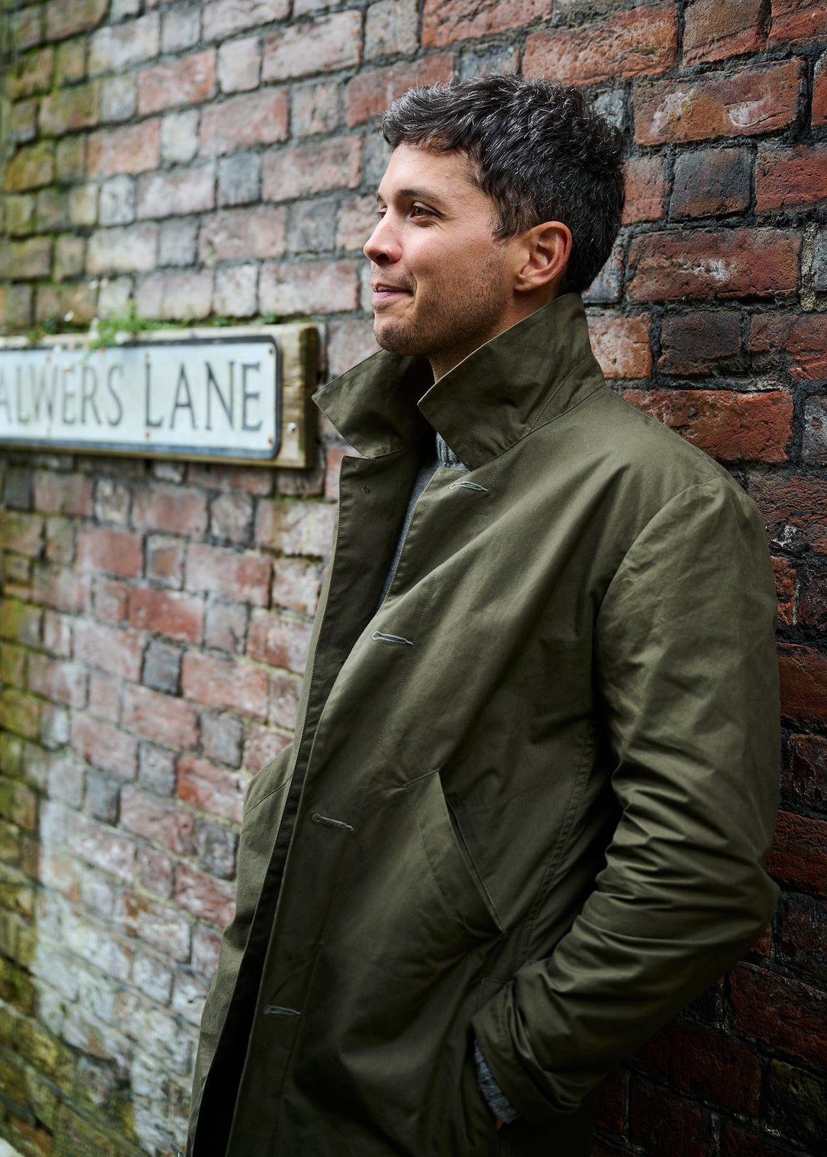 A man leaning againsta brick wall wearing a dark olive colour cotton duster coat