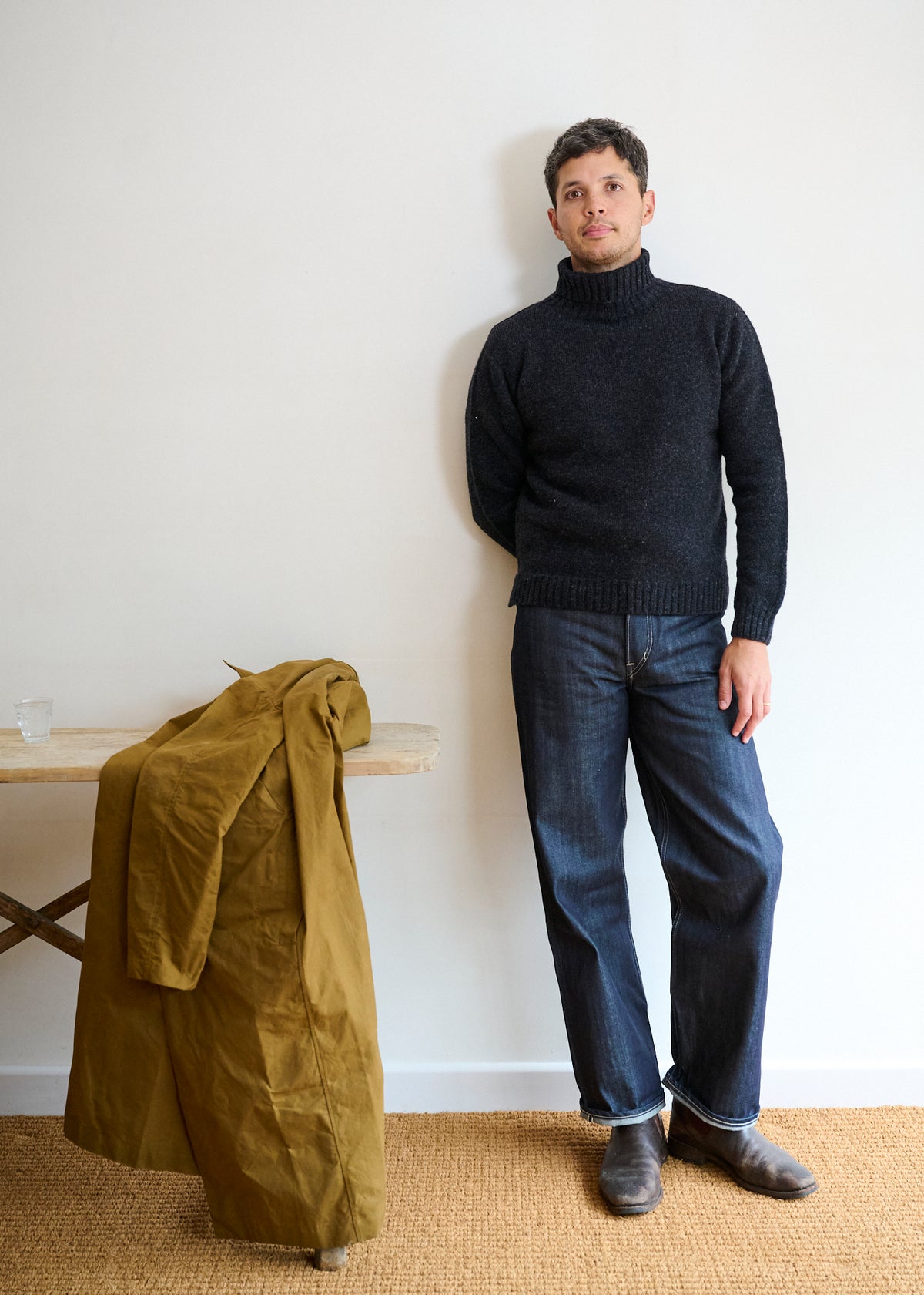 A man wearing a charcoal gray roll neck lambswool plain knit jumper and blue jeans, standing next to a green jacket on a stand.