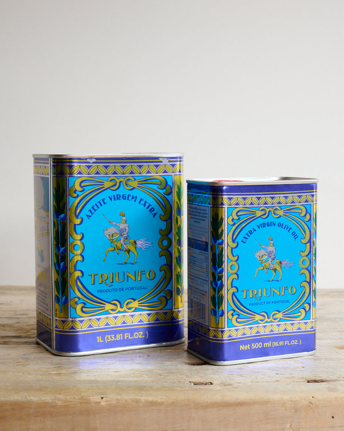 Two traditional tin cans of Triunfo Portuguese Extra Virgin Olive Oil on a wooden surface.