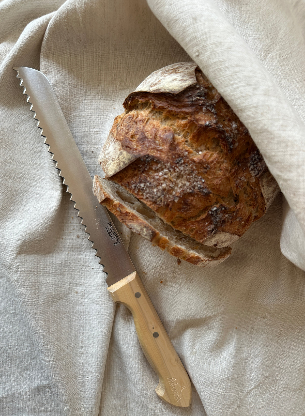 Stainless steel bread knife with a light wood handle next to a round loaf of sourdough bread on a linen cloth