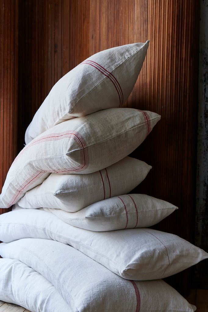 A stack of beige linen grain sack cushions with red stripes, placed against a wooden backdrop.