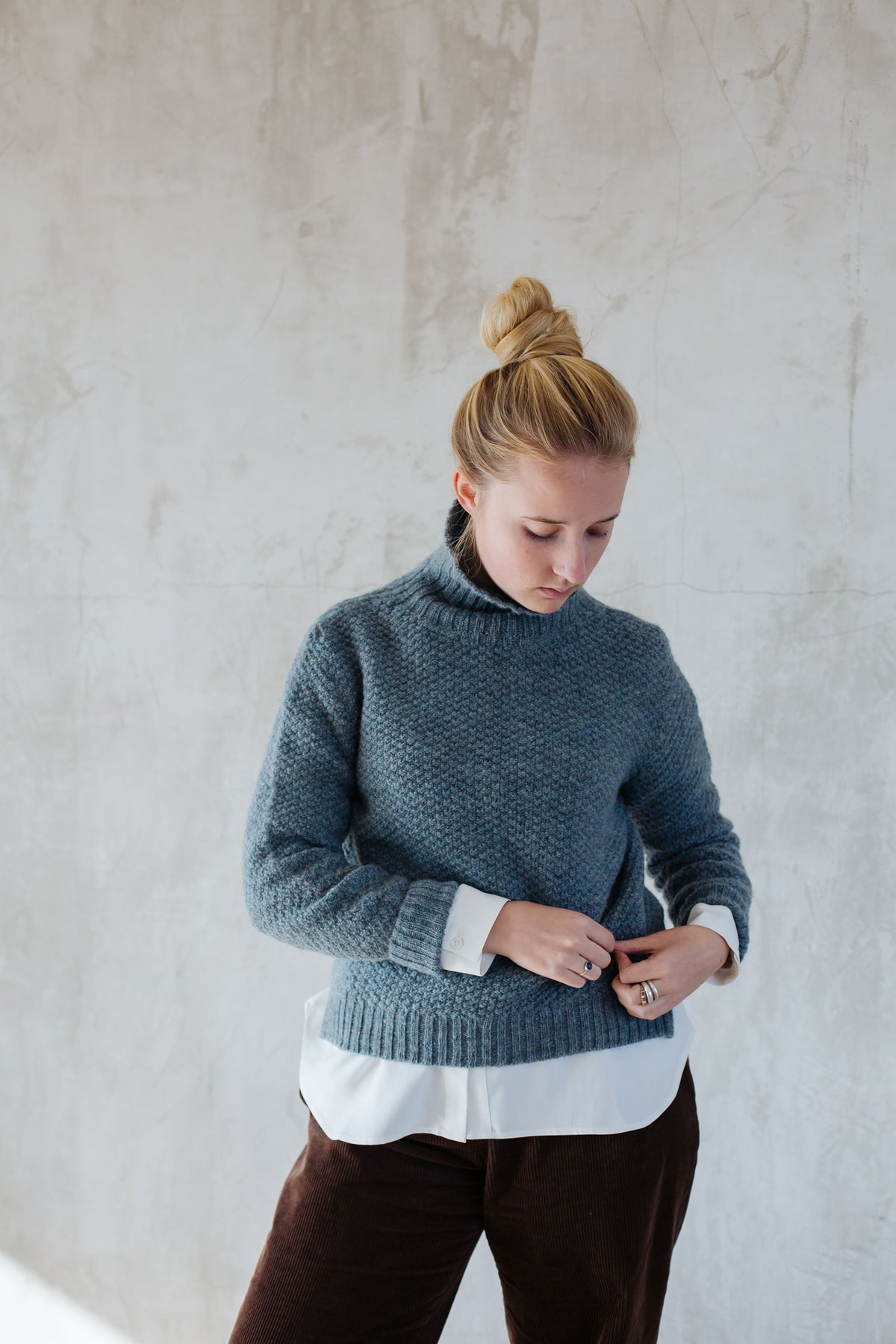 A woman wearing a graphite gray moss stitch lambswool jumper with a funnel neck, slit side detail, and a longer back length.